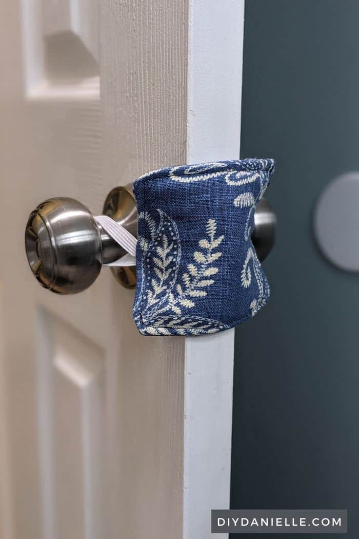 DIY Door Silencer. Picture of pretty blue floral fabric square, attached by elastic around the back and front of a door knob. This blocks the closing piece of the door so it's quieter to close.