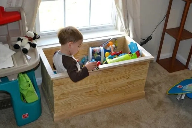 Toddler sitting in a toy chest full of toys.