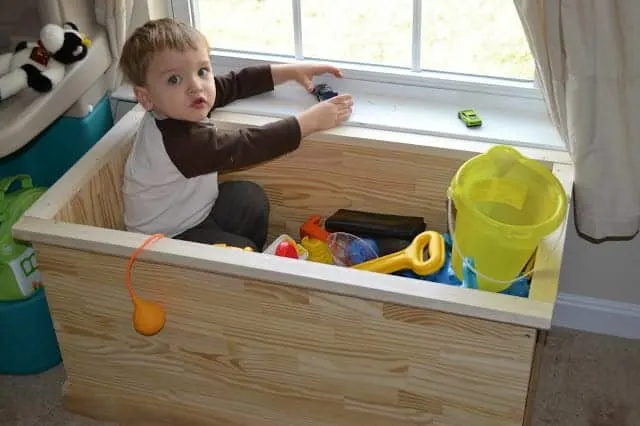 Toddler sitting in a toy chest full of toys with toy cars on the windowsill. 