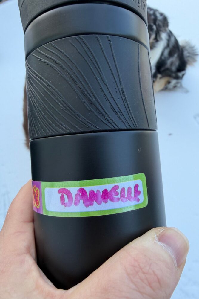 Mabels Labels on a Contigo insulated water bottle. Label has a butterfly on the left side, pink and green coloring, and pink handwriting with a dry erase marker that says Danielle. Water bottle is black.