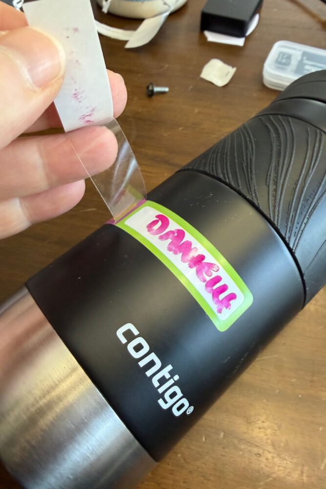 Mabels Labels on a Contigo insulated water bottle. Process of peeling off the backing from the clear part of the label.

Label has pink and green coloring, and pink handwriting with a dry erase marker that says Danielle. Water bottle is black.