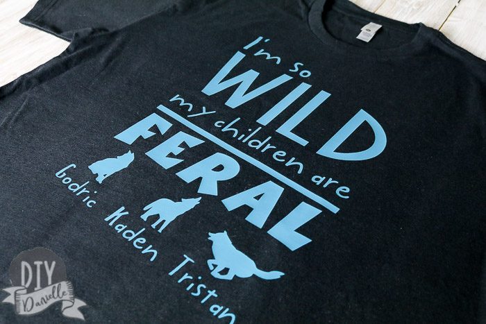 Black shirt with blue lettering that says "I'm so wild my children are feral" with three wolves and the kids names underneath.
