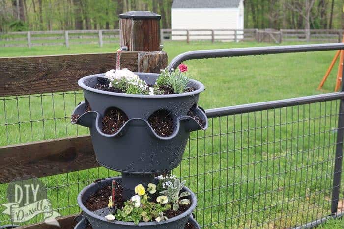 Large, three tier self watering planter with flowers inside it. Pictured by our backyard fence.