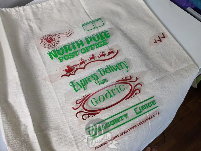 DIY Personalized Santa Bag: Individual pieces of HTV laid out in order to create the entire red and green design. Design says "North Pole Post Office, Express Delivery for Godric" with a naughty or nice check box.