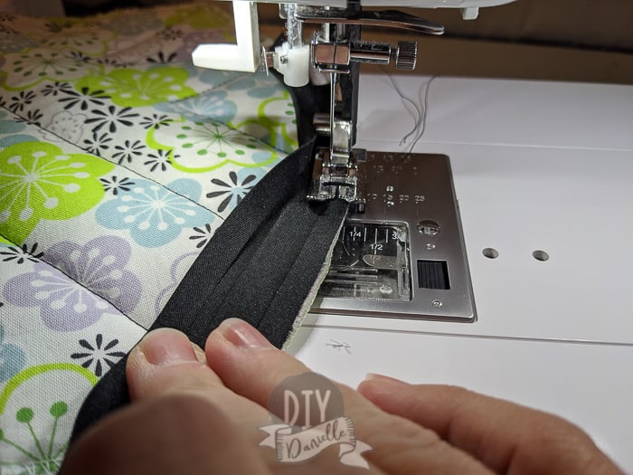 Double fold bias tape being sewn onto a pot holder that has purple, green and blue floral print. 