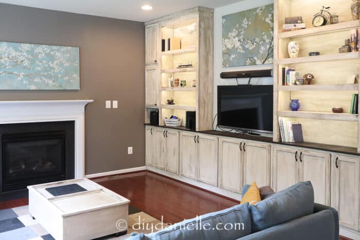 DIY Entertainment Center that's built-in with under cabinet lighting, bookshelves, and storage space. Distressed paint job for rustic/farmhouse look. TV wall mounted between the bookshelves.