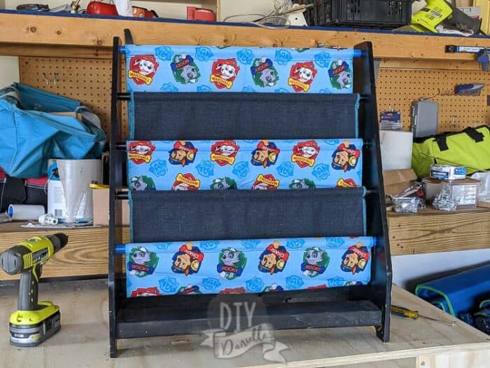 Repaired sling bookshelf sitting on top of a toolbench with tools nearby. Paw Patrol and matching dark blue fabric for the slings.