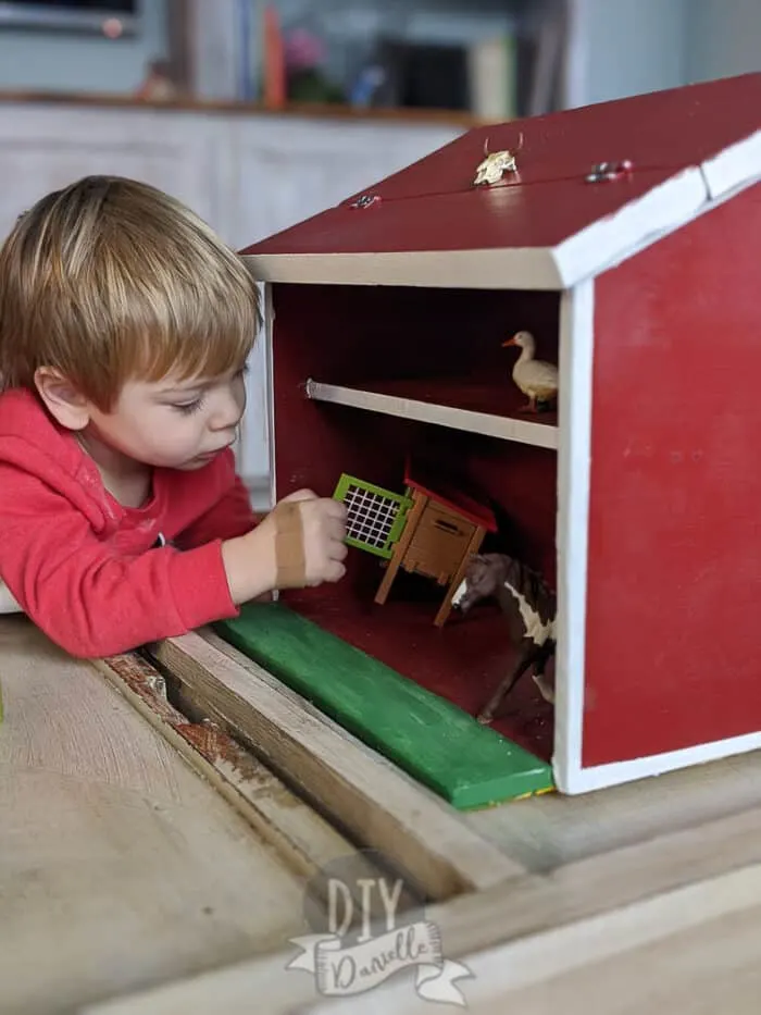 Little boy playing with a wood toy barn painted red with white trim. Farm animals inside the barn include a duck, rabbit hutch, and a horse.