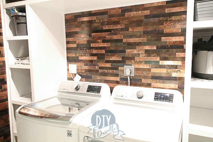 Aspect Backsplash in Metal and Aged Copper behind the washing machine and dryer. White shelves framing the wash area.