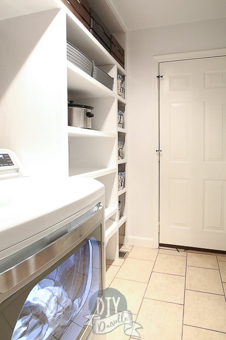 DIY laundry room shelving and dryer pictured, looking from laundry sink to exit door. Door stops on the door to the garage keep the door from denting the handmade shelving. 