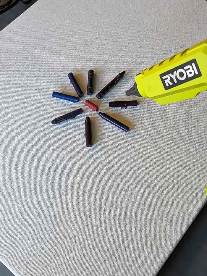 Using my Ryobi glue gun to glue the crayons (with labels peeled off) down to the canvas. The red piece of crayon is in the middle with the blue and purple pieces in a starburst shape around it.