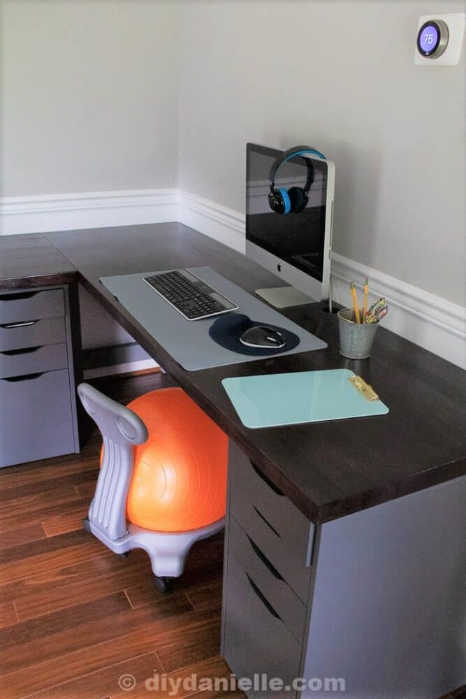 iMac with headphones on it, birch butcherblock with dark cherry stain, a yoga ball chair, clipboard, gray container of school supplies, and keyboard/mouse on top of a desk protector mat. This is the homeschooling station for one of my sons. The full desk is an L shaped corner desk and big enough for two kids.