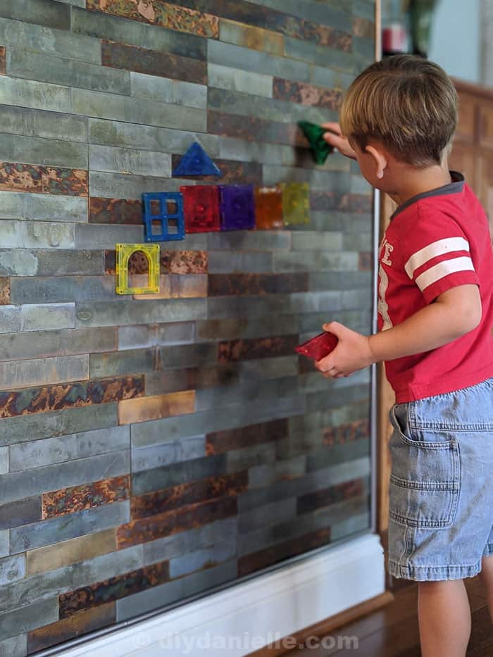 Distressed metal stick on tiles with toddler boy placing magnetiles on the wall.