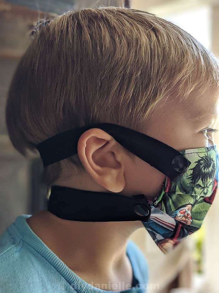 3 year old boy with blond hair wearing a super hero mask with elastic straps that snap on around the back of his head and neck. 