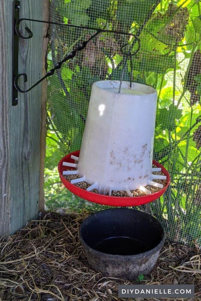 Chicken feeder hanging off a post in the chicken run from a plant bracket. Bucket underneath to catch feed that spills.