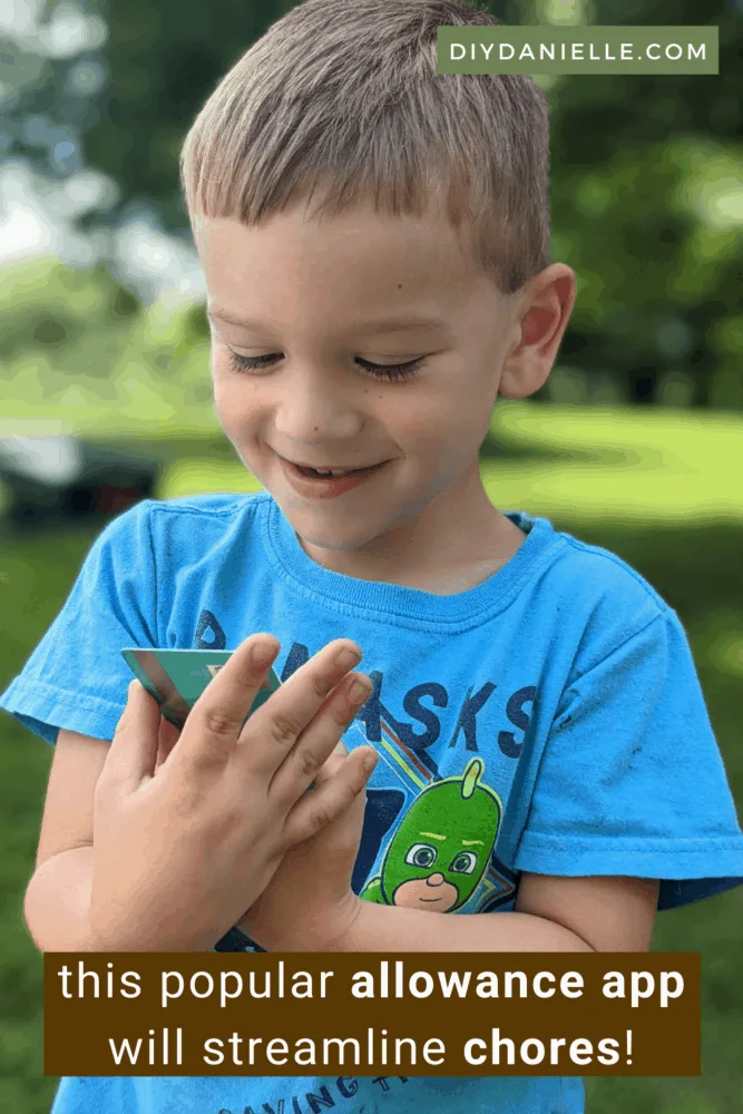 This popular allowance app for kids will streamline chores. Photo of a blond toddler in a blue shirt holding a debit card in his hands and looking at it with a smile on his face.
