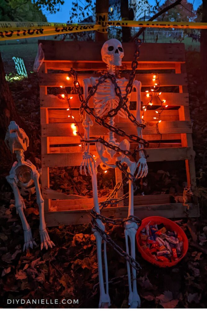 Skeleton chained to a pallet with red lighting behind him., a skeleton dog beside him and a bowl of candy on the ground.