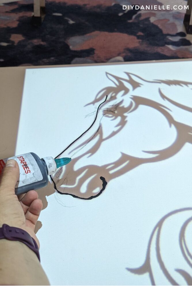 An image of a horse head is projected onto a large white canvas. Someone's hand is holding a glue bottle filled with a mix of glue and black acrylic paint over the design, tracing the design with the black glue. 