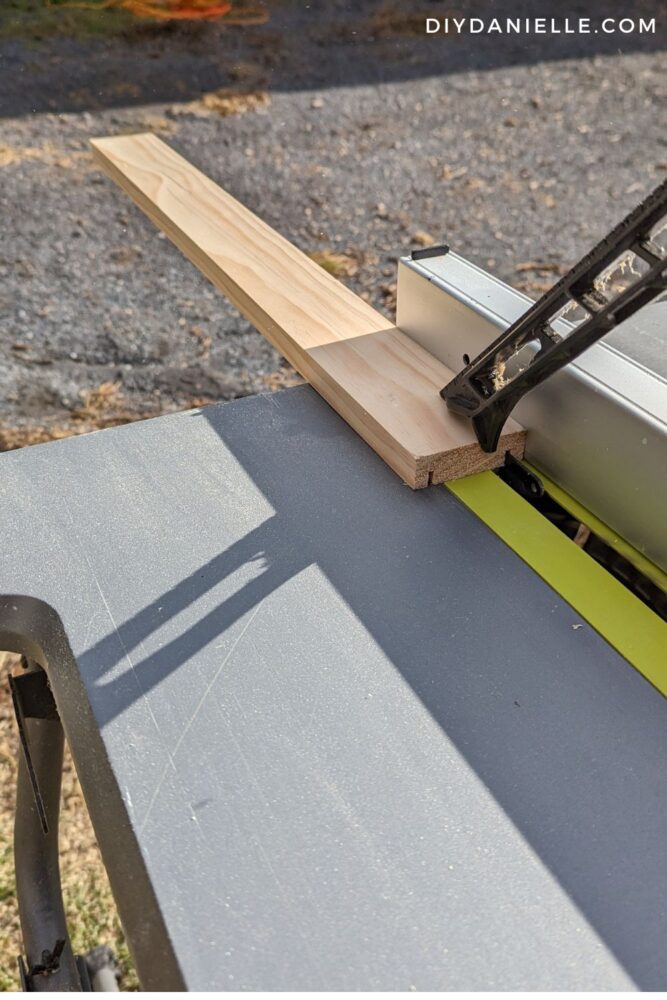 Photo of a piece of 1x3 wood being groove cut on a table saw. Person (not shown) is using a tool to push the wood through.