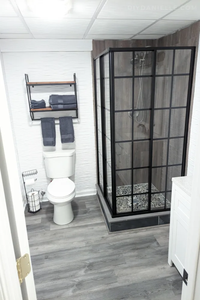 Photo of the finished basement bathroom. The white walls and ceiling are offset with darker tones in the gray and brown flooring and shower tiles. The square shower has framed black glass doors.