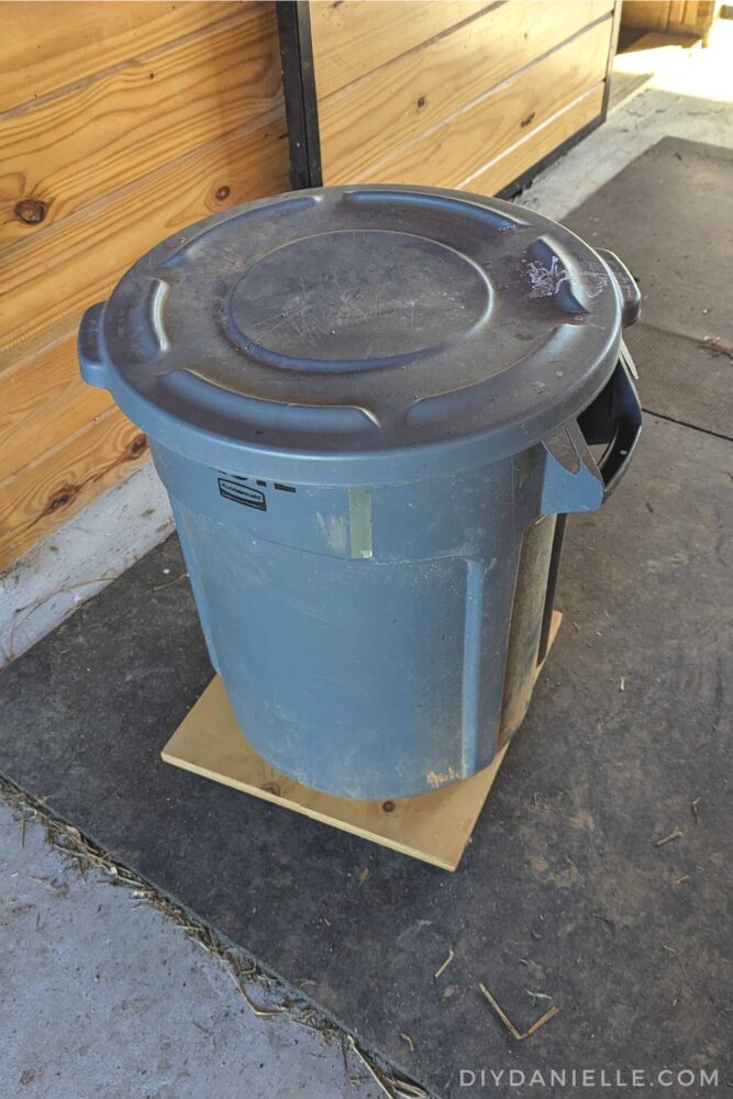 Feed bin on wheels, piece of plywood with the plastic garbage can on top. Top down view.