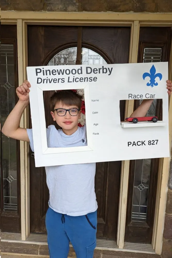 Pinewood Derby Drivers Licensed DIY Peep Board: This white board has two cut outs, one for the child to hold up to their face and another to display their Pinewood Derby race car. The parents can edit in the child's name, date, age, and rank. The pack number is at the bottom right. There's a blue Scouts logo at the top.