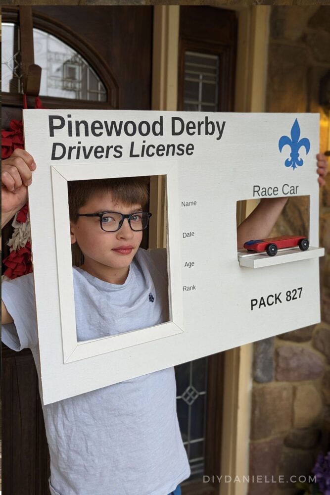 Pinewood Derby Drivers Licensed DIY Peep Board: This white board has two cut outs, one for the child to hold up to their face and another to display their Pinewood Derby race car. The parents can edit in the child's name, date, age, and rank. The pack number is at the bottom right. There's a blue Scouts logo at the top. An 11 year old Cub Scout is holding the photo board with his red car displayed.