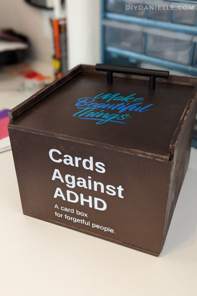 DIY Card Box painted metallic brown. The top lid has a pull attached so you can pull it and slide the lid off. The top says "Make beautiful things" in two tones of blue permanent Cricut vinyl. The front of the box says "Cards against ADHD: A card box for forgetful people."
