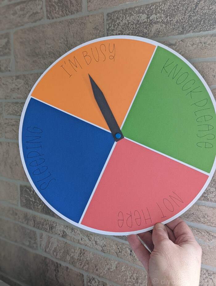 Availability clock made from cardstock and Smart Sticker Cardstock. The white circle is divided into four quadrants that  say "Knock Please" "I'm Busy" "Not here" and "Sleeping" and there's a black arrow pointer hooked to the paper sign using a small paper fastener. 
