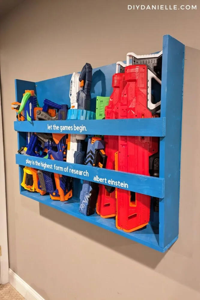 Nerf gun storage rack on the wall, full of Nerf guns and bows. This is a Nerf gun rack that I built for the wall in our playroom. It says "let the games begin" in Cricut vinyl on the top piece of wood, then "Play is the highest form of research. Albert Einstein" on the second piece of wood. The wall mounted shelf is painted teal and it fits quite a few Nerf guns. I think there are 7 or more guns on this shelf in the picture.
