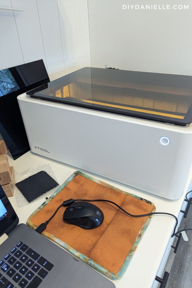 Review of the xTool M1 10W Laser Cutter Machine: Image shows the machine setup and connected via a USB cord to a laptop. There is a computer mouse on a mouse pad, and a blank coaster waiting to be engraved.