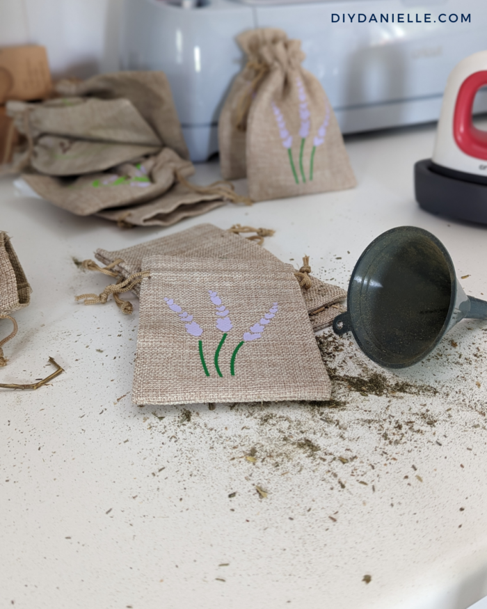 Photo of 5 small burlap bags filled with dried lavender. The bags are customized with a lavender design in iron-on vinyl. These bags haven't been filled yet but there's a funnel next to them and dried lavender on the table.

These bags are cute gifts for roommates and hosts while traveling because they don't take up much space in luggage.