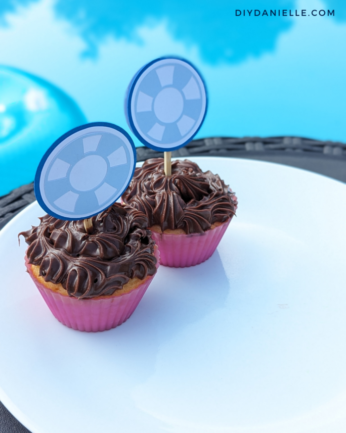 Pool Party Idea: Cricut Made Cupcake Toppers in the shape of pool floats, photograph in vanilla cupcakes with chocolate frosting with a pool in background.