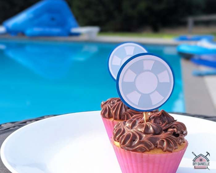DIY Cupcake Toppers that look like a light blue and white pool float. They are stuck in some vanilla cupcakes with chocolate frosting. In the background is a pool and some pool floats which are blurred out. 

These pool party cupcake toppers are made with cardstock and Cricut's SmartSticker Cardstock and have a skewer in between the layers to stick into the cupcake.