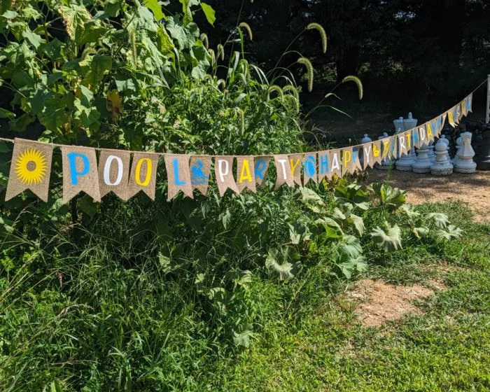 Photo of a burlap sign with flags that say "Pool Party Happy Birthday Tristan"... the colors alternate between blue, white and yellow, and there are swimming/sunshine related images separating each word. In the background is a giant chess set and some green plants (weeds with a volunteer pumpkin plant).