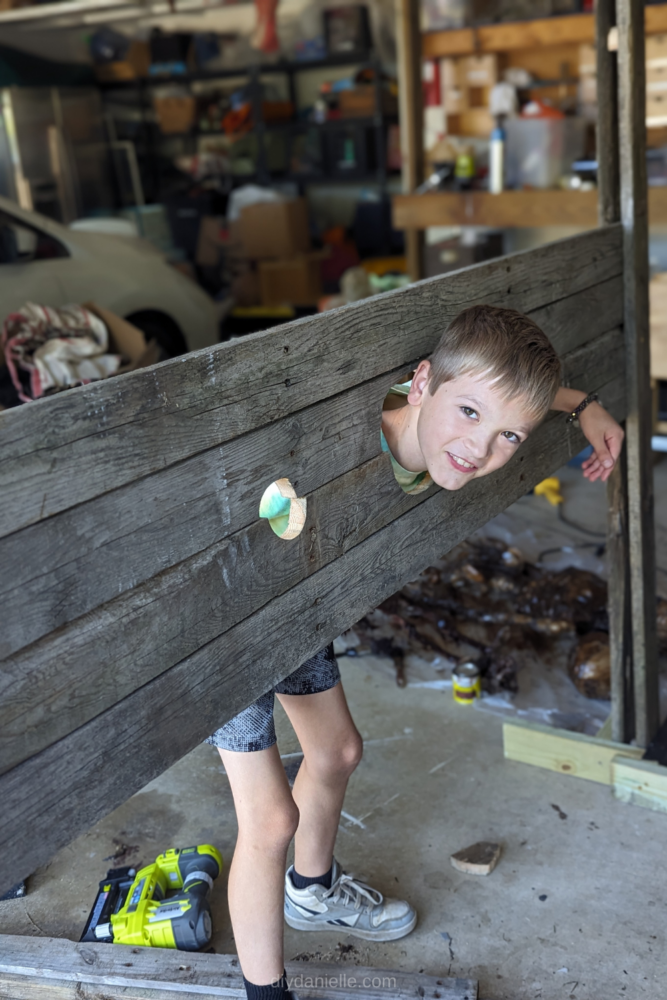 DIY Stocks with a smiling 9 year old boy with blond hair and brown eyes inside them, with one hand out. The hand spots for the stocks are a big large so he was able to get out easily. 