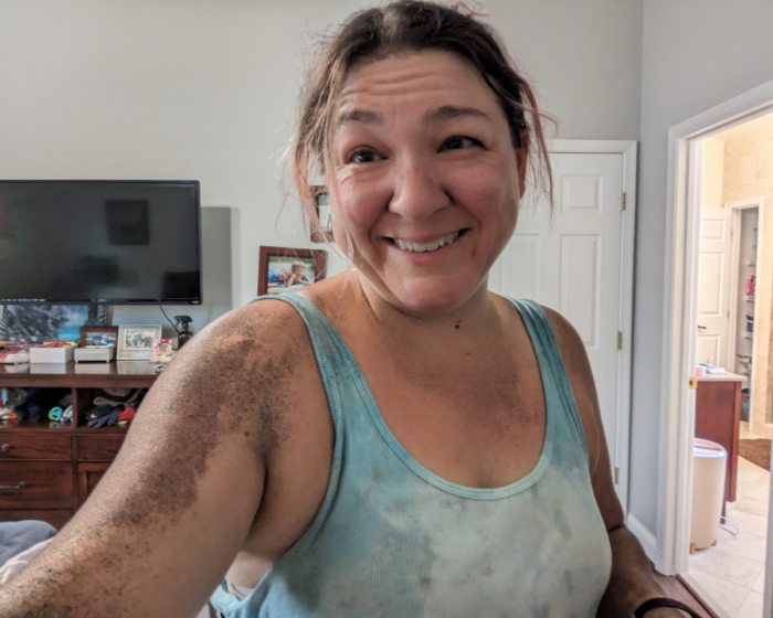 Photo of myself after I spray painted our new goat and sheep barn black. I'm covered in black spray paint... which looks like I rolled in the mud. 

Photo of a white woman in her 40s in a blue tie dye shirt with black spray paint on her from overspray from using an airless paint sprayer.