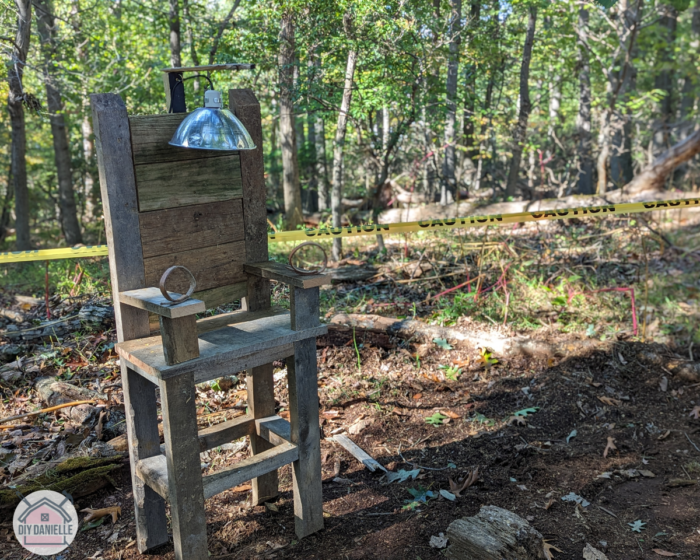 Photo of the electric chair that I built for Halloween decorations this year. The chair is setup in the woods with yellow caution tape behind it. There's an electric headpiece, arm straps and the wood is aged.