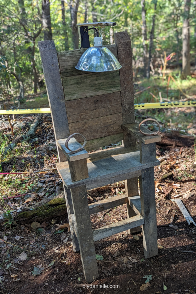How to make a DIY electric chair prop. This outdoor Halloween decoration is made out of lots of aged scrap wood! 

Image: Photo of a fake electric chair setup in the woods with caution tape behind it. There are closed straps for holding the arms down, and an electric 'hat' made from a chicken brooder heat lamp for the heat piece. The wood is all old and looks aged.