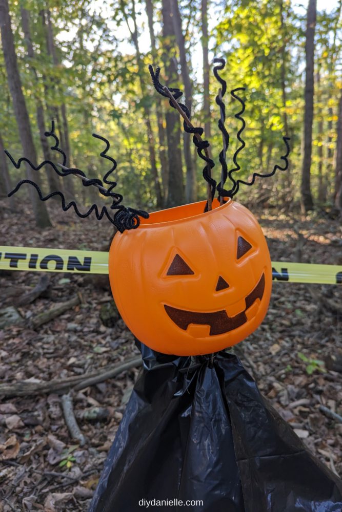 Black plastic bag flipped upside down over a tomato cage. There is a plastic pumpkin on top. The metal ends of the tomato cage are covered with curled black pipe cleaners to appear like curly hair.