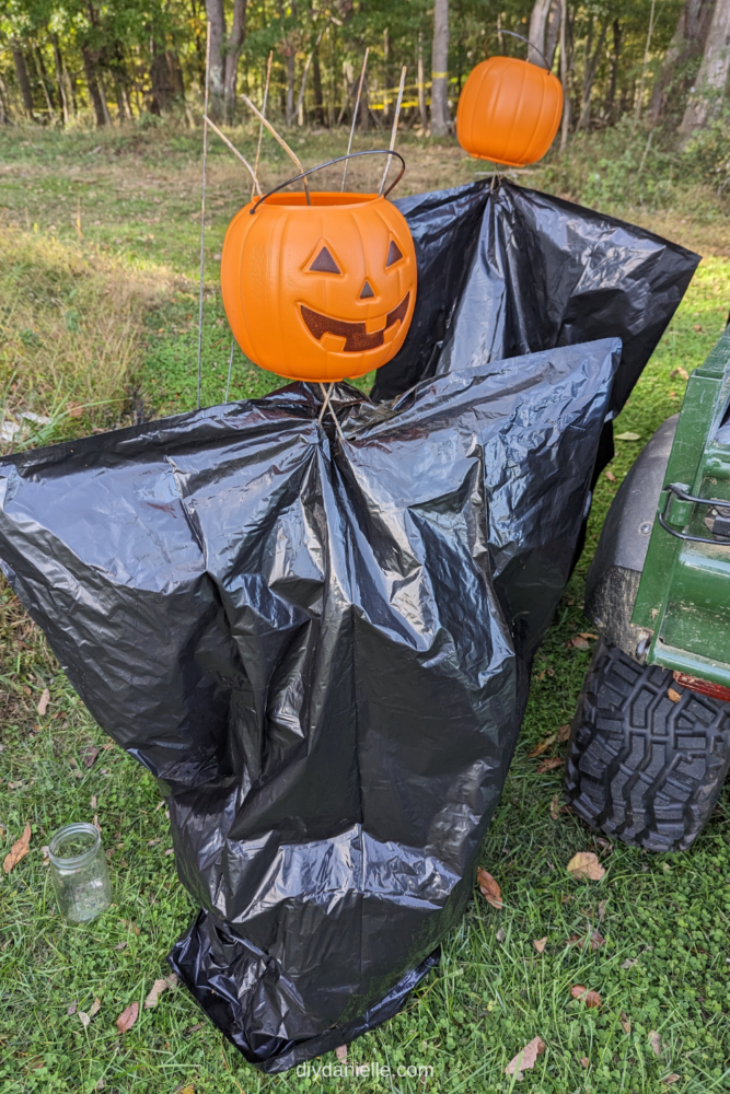 Black plastic bag flipped upside down over a tomato cage. There is a plastic pumpkin on top.