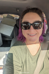 White woman with pink and brown hair and sunglasses seat belted in her car with white foam blocks in the back.