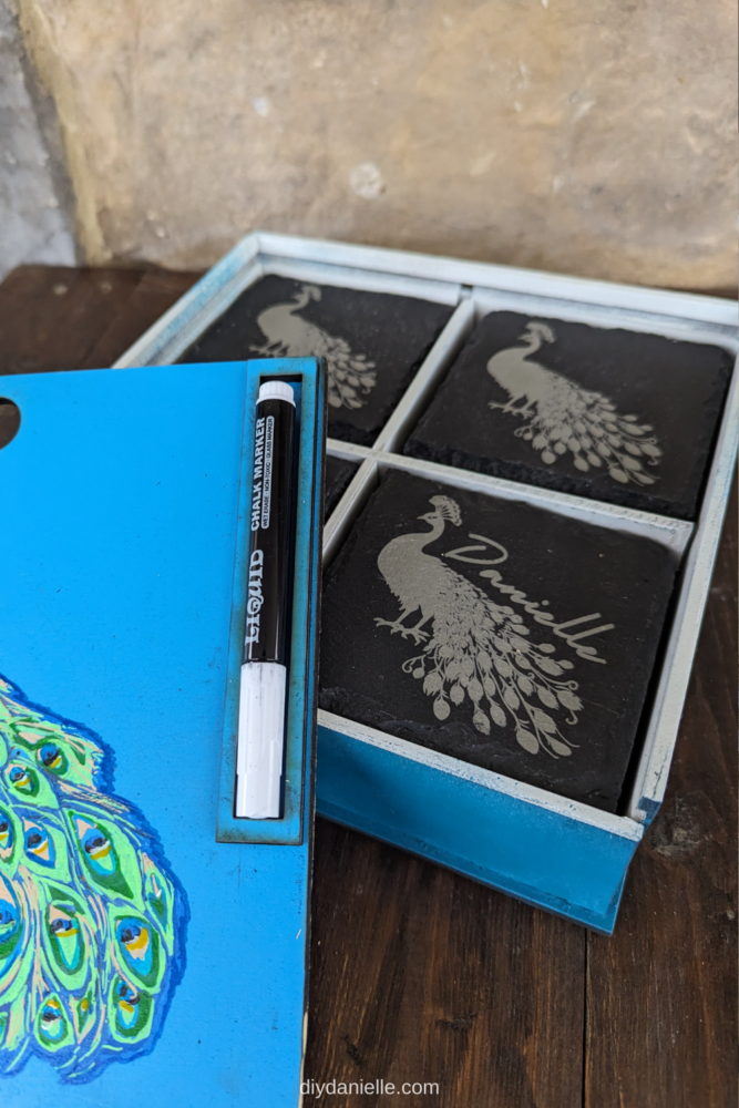 DIY Coaster Box with a Peacock on the cover. The cover slides on and off easily for accessing your coasters. 

This photo shows the open box with the lid placed to the side. You can see the coasters engraved with peacocks, and some are engraved with the person's name above the peacock.

There's a spot to place a chalk marker on the top. The chalk marker allows you to write names on your coasters to mark where people are seated.