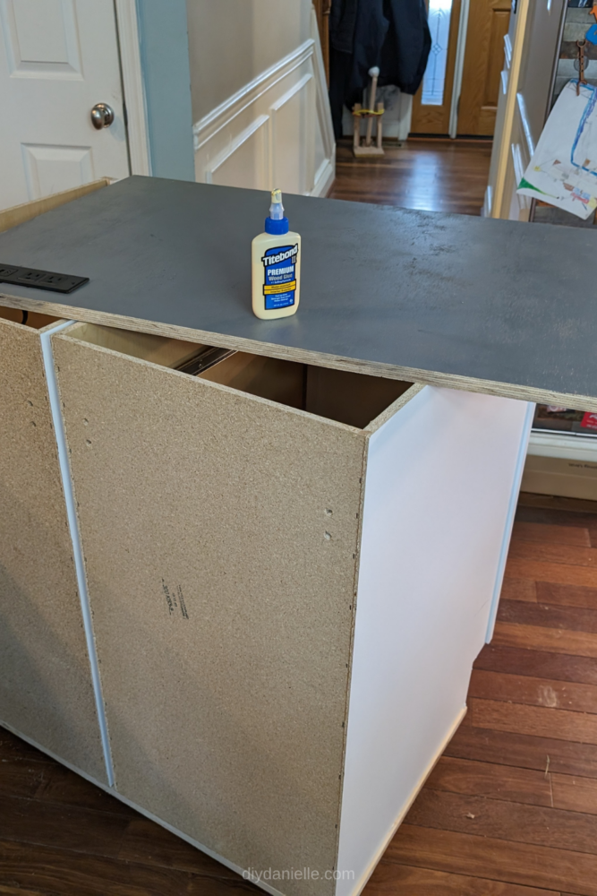 Use wood glue to attach the painted plywood countertop to the base cabinets on the microwave cart before securing it with nails.