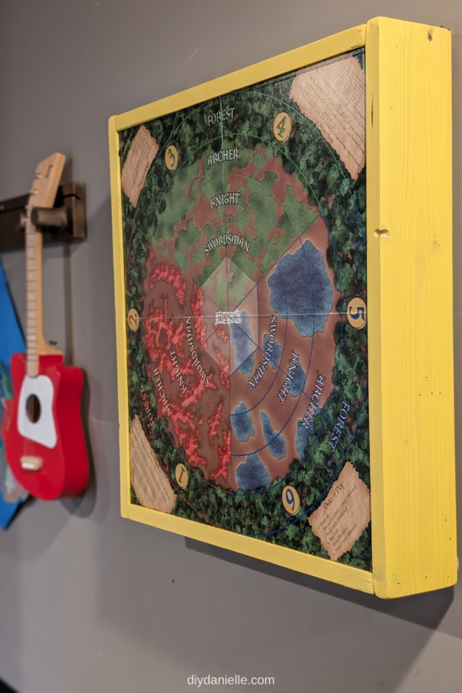 The DIY board game display hangs on the wall next and features the game board on the front and a colorful, yellow wooden frame around the sides.