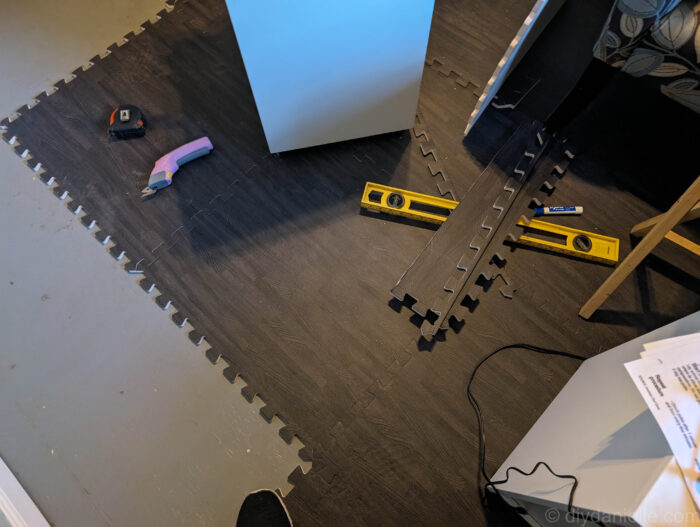 Foam floor tile in dark gray wood grain with the trim pieces on it. This photo was taken during the installation process and shows scraps, electric scissors, a measuring tape, and a straight edge ruler for drawing the lines with the blue dry erase marker. 