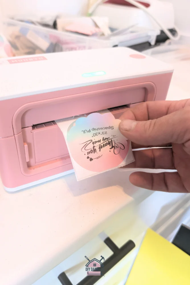 Pink Munbyn compact label printer that uses thermal printing technology to print labels. The printer is printing on a flower shaped label with rainbow colors. a person's fingers are grabbing the label as it comes out of the machine. The label text says "Thank you very much! 23x30" Spaceship PUL." These are labels for some fabric that I'm selling.