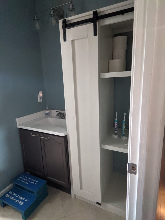 DIY Bathroom Cabinet painted white with sliding doors that have barn door style hinges. Next to a small sink with a step stool for kids to reach the sink to brush their teeth. Toilet paper and toothbrushes are stored on the shelves.