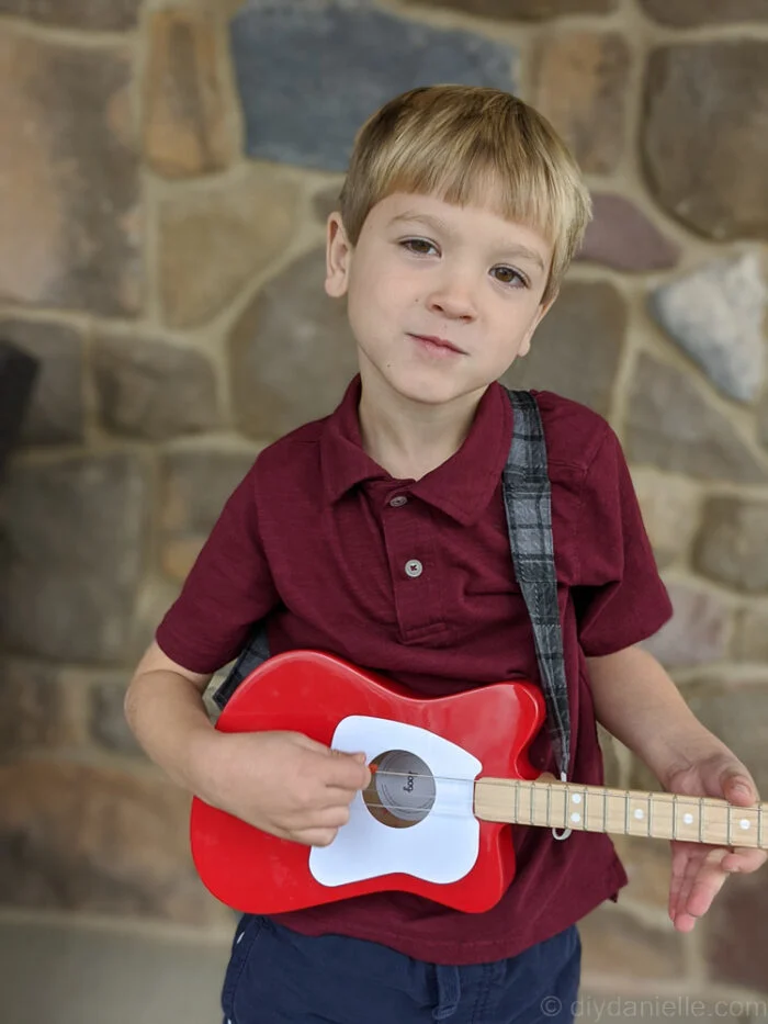 Young boy facing forward, holding a guitar and wearing a custom fabric guitar strap, smiling at the camera.