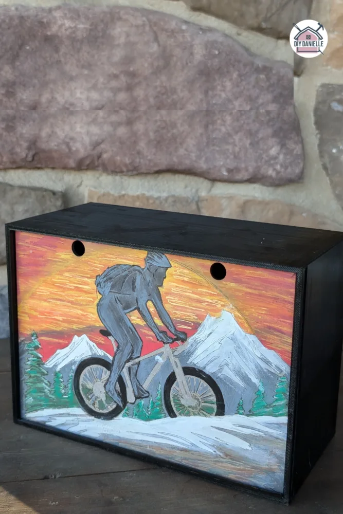DIY Memory Box: The base is 3D print using black PETG filament, and the sliding lid is laser cut and engraved.
The orange side of the lid (the side showing in the photo) has a man riding a mountain bike with a sunset and mountains and trees in the background. There's some snow on the ground. The man is more of a silhouette in tones of gray.